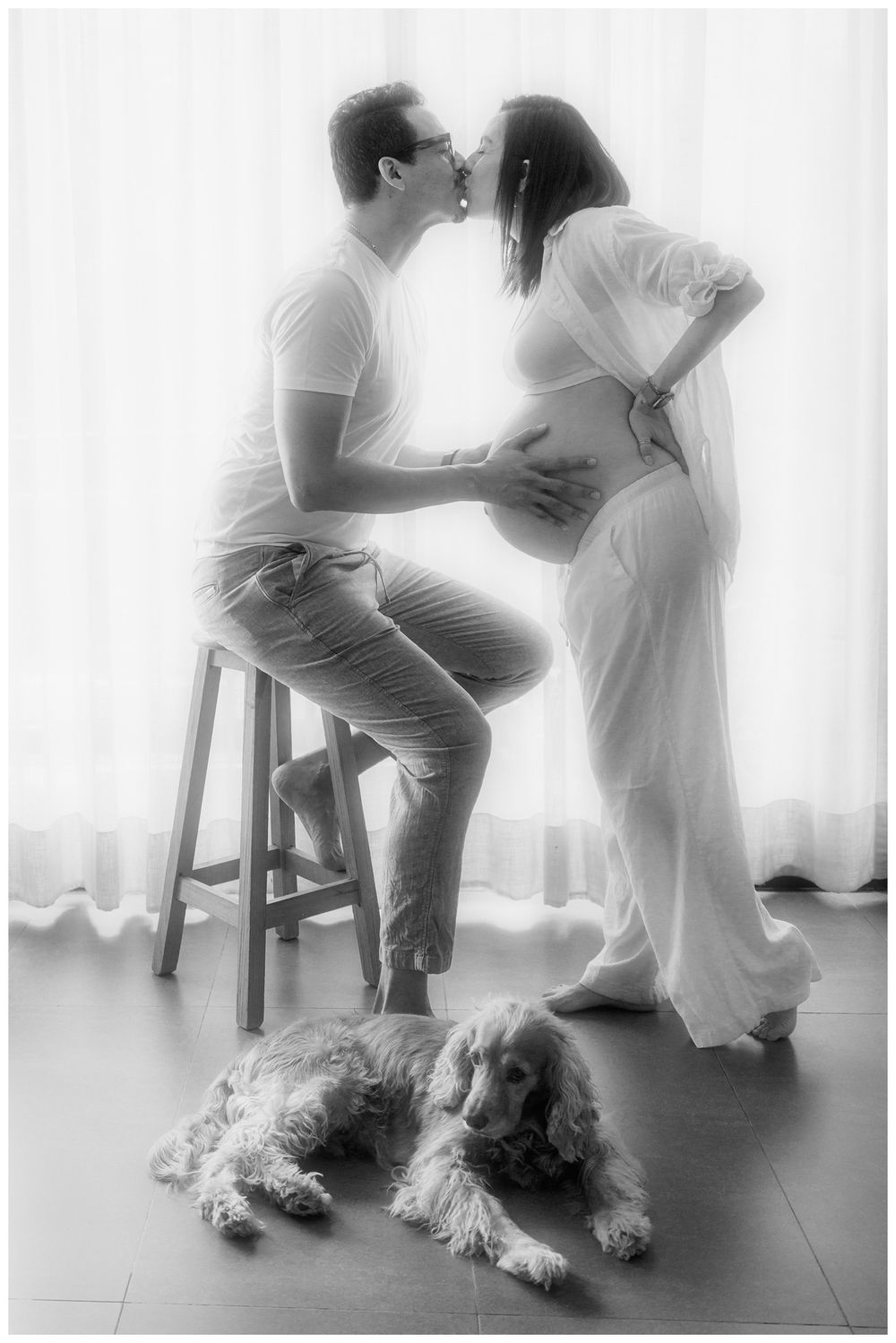Black and white maternity photo of kissing couple with dog, pregnant woman in white dress, man on stool by sheer curtains.