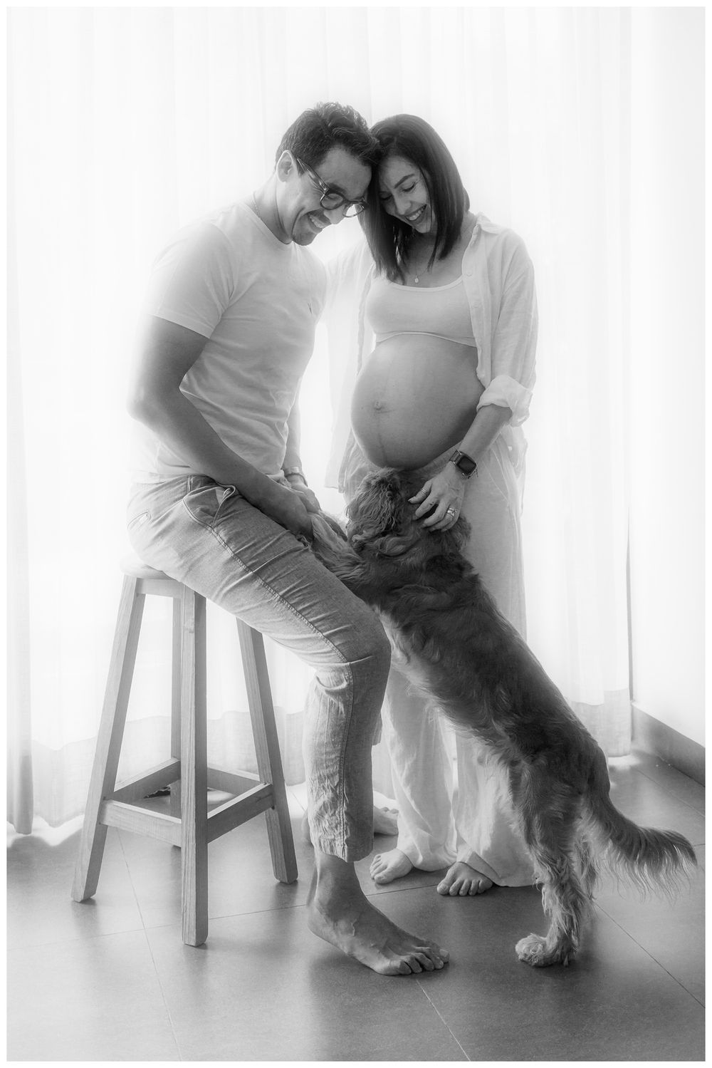 Black and white maternity photo of couple with dog standing on hind legs touching pregnant belly.