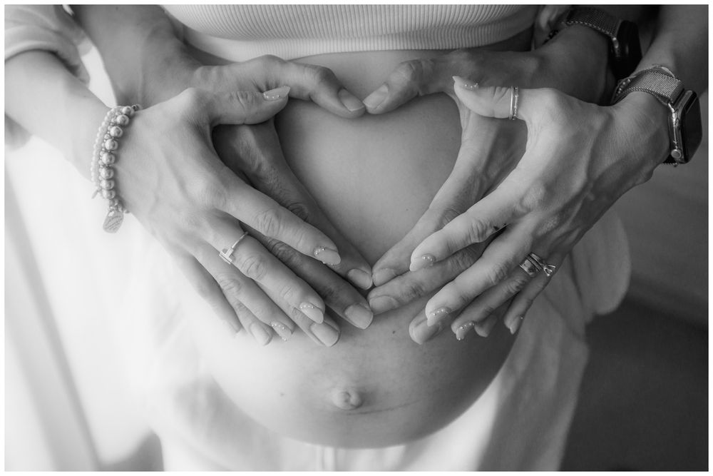 Black and white maternity photo of couple forming heart shape with hands on pregnant belly, wearing rings and bracelets.