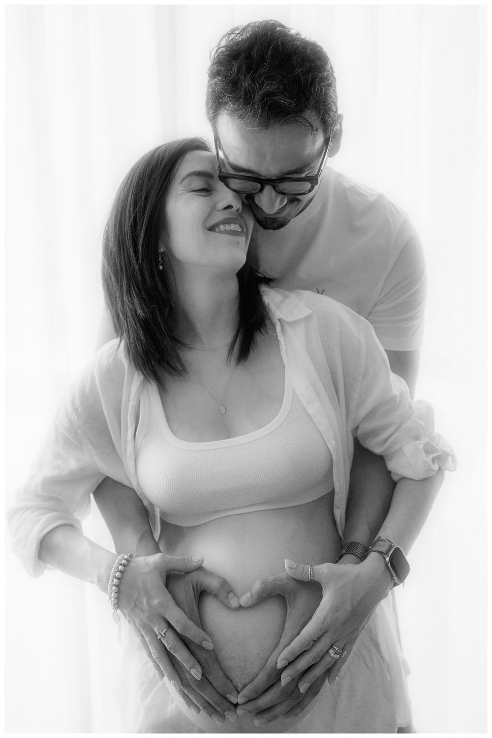Couple forming heart shape with hands on pregnant belly in black and white maternity photo.
