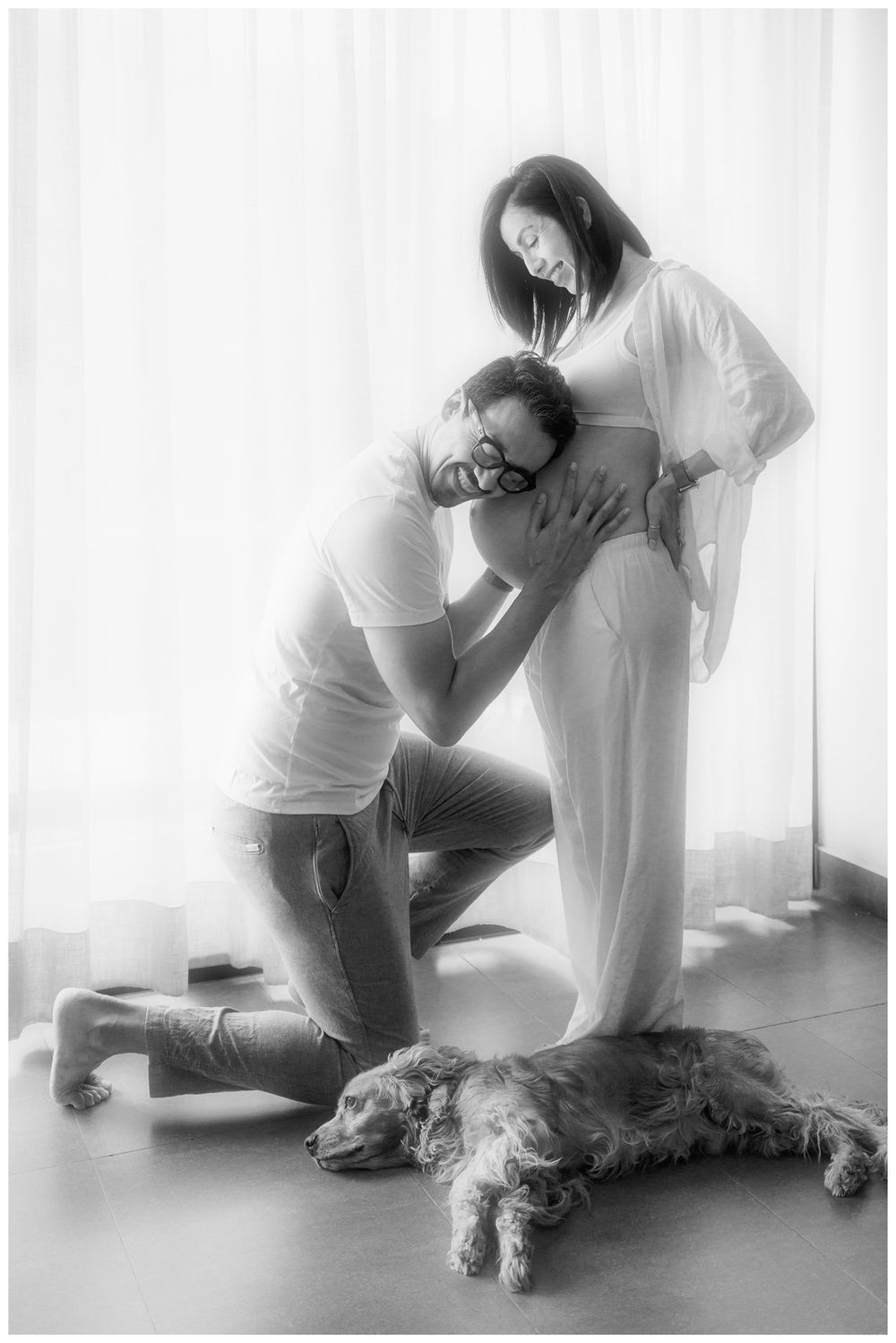 Man kneeling to listen to pregnant partners belly with dog lying nearby in B&W photo.
