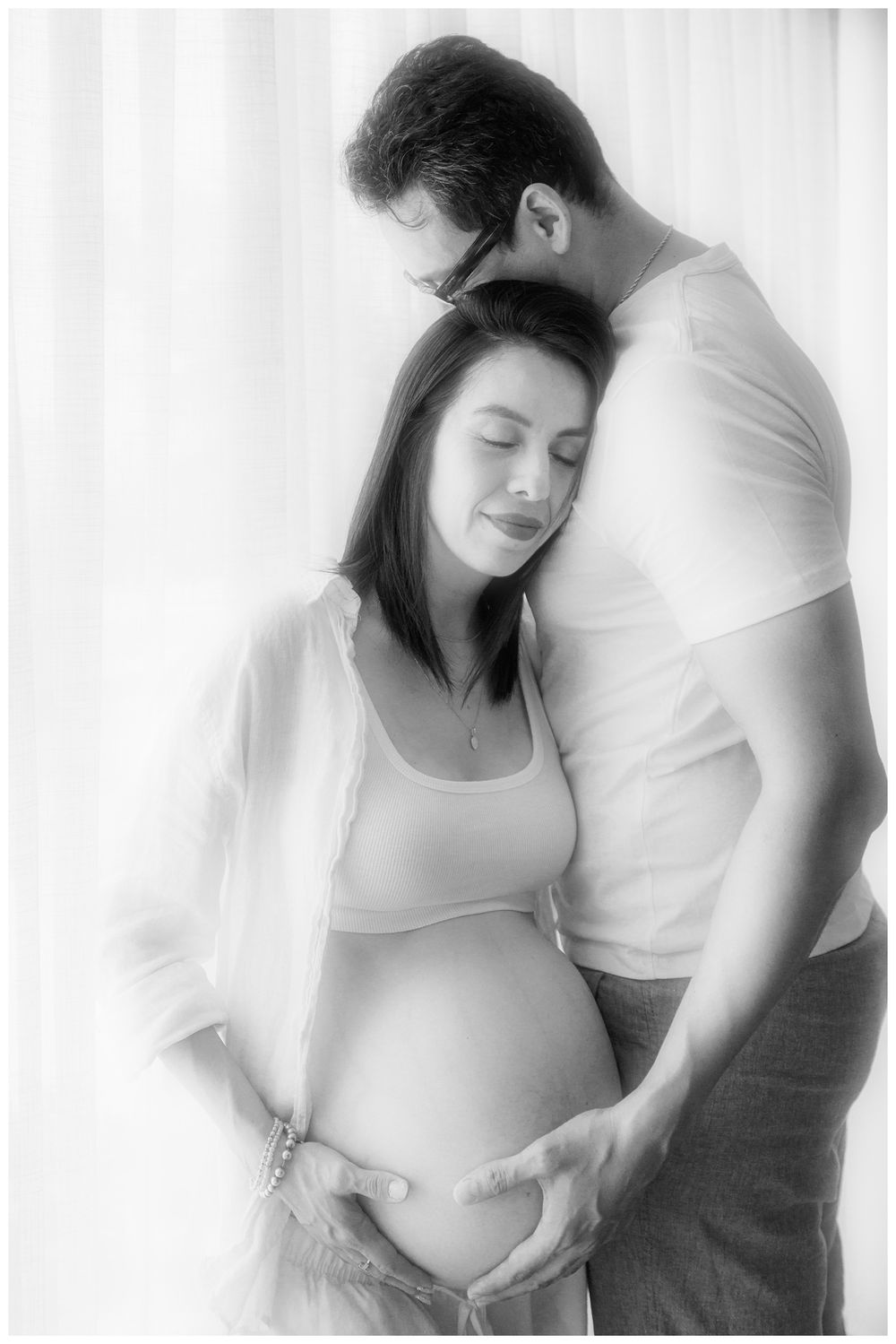 Tender black and white maternity portrait of man embracing pregnant woman from behind near window.