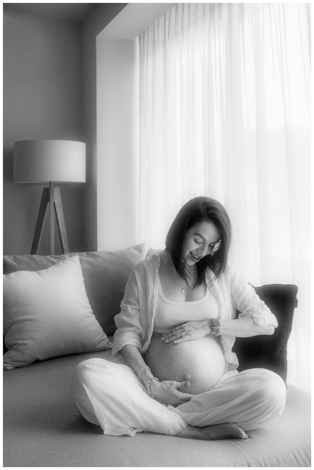 Black and white photo of a smiling pregnant woman sitting cross-legged on a sofa, cradling her baby bump.