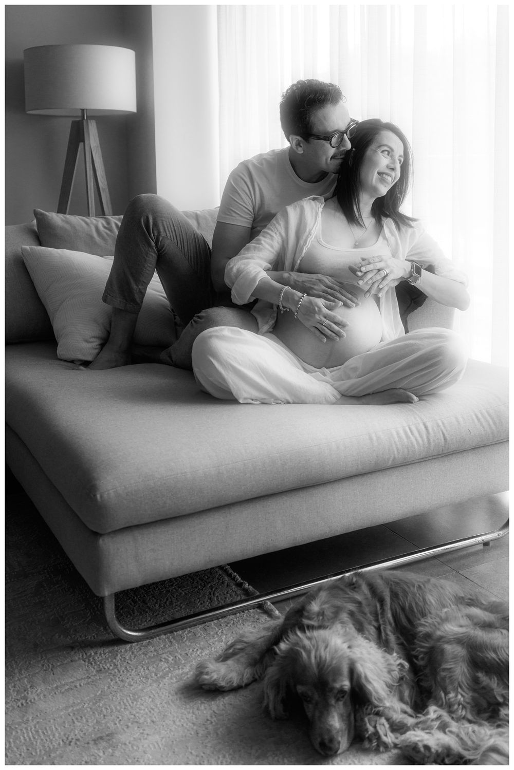 Black and white maternity photo of couple sitting on sofa with dog resting on floor in cozy home setting.