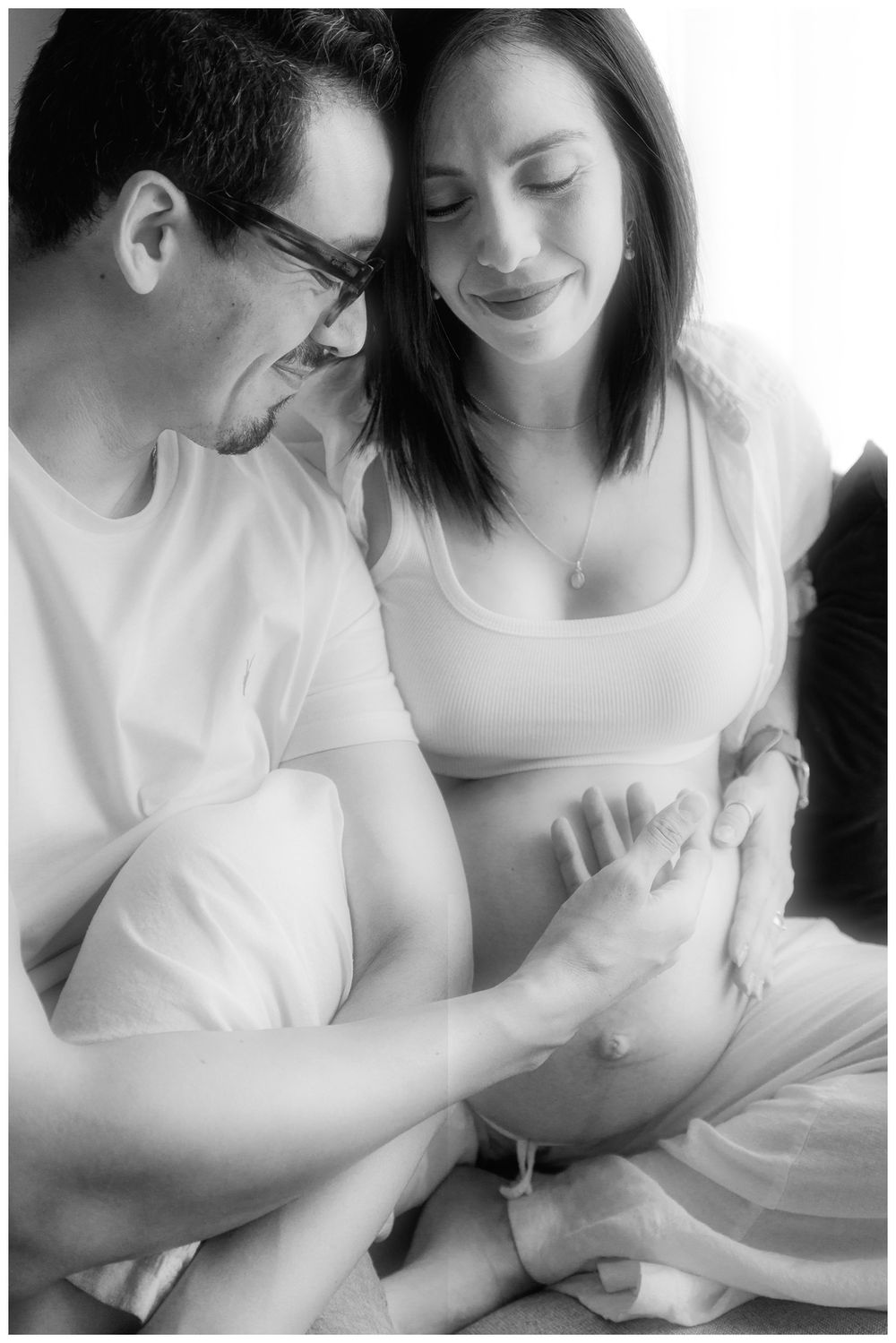 Black and white photo of couple smiling and holding pregnant belly together in intimate moment.