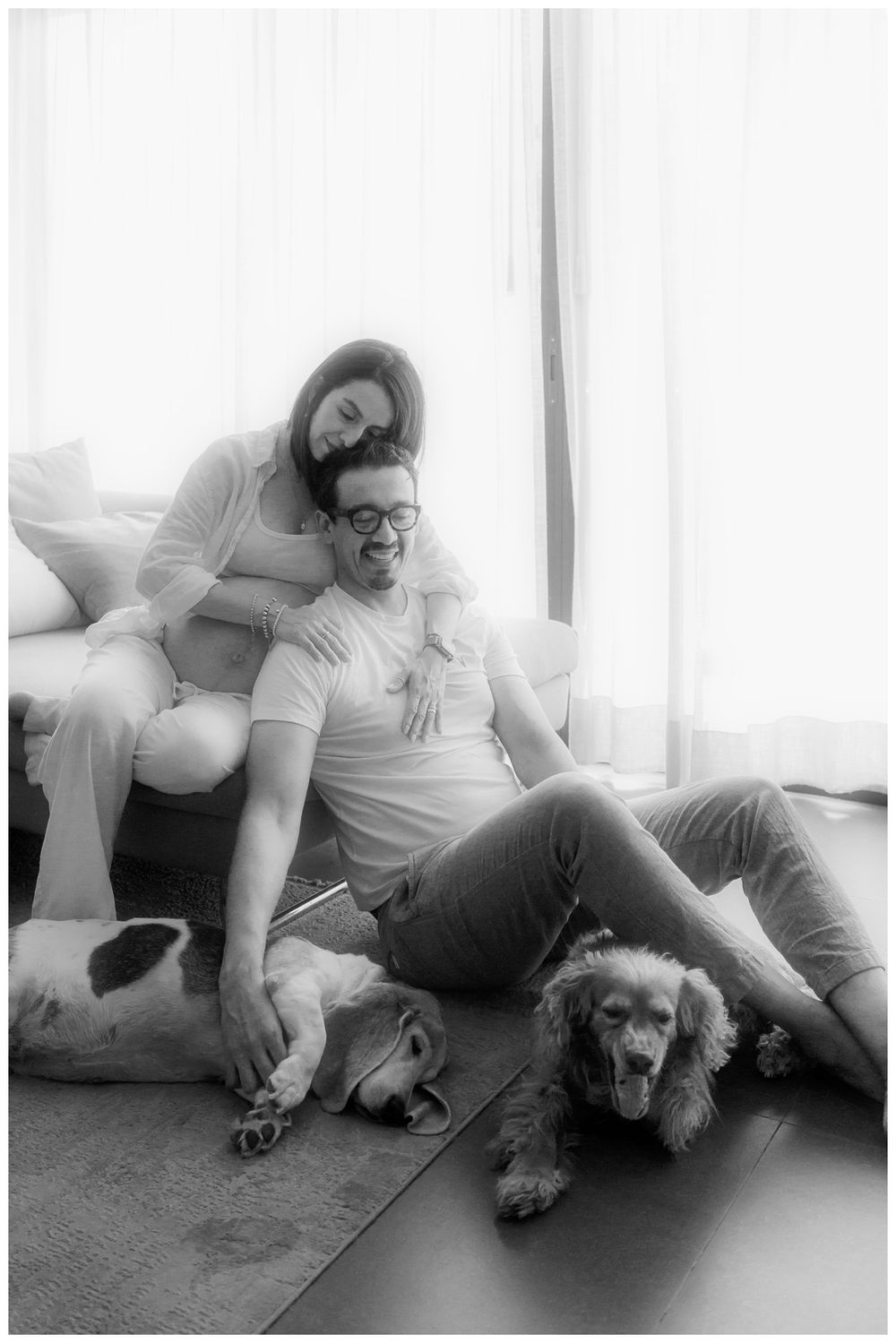Black and white photo of a couple relaxing on the floor with two dogs, smiling warmly in a bright living room.