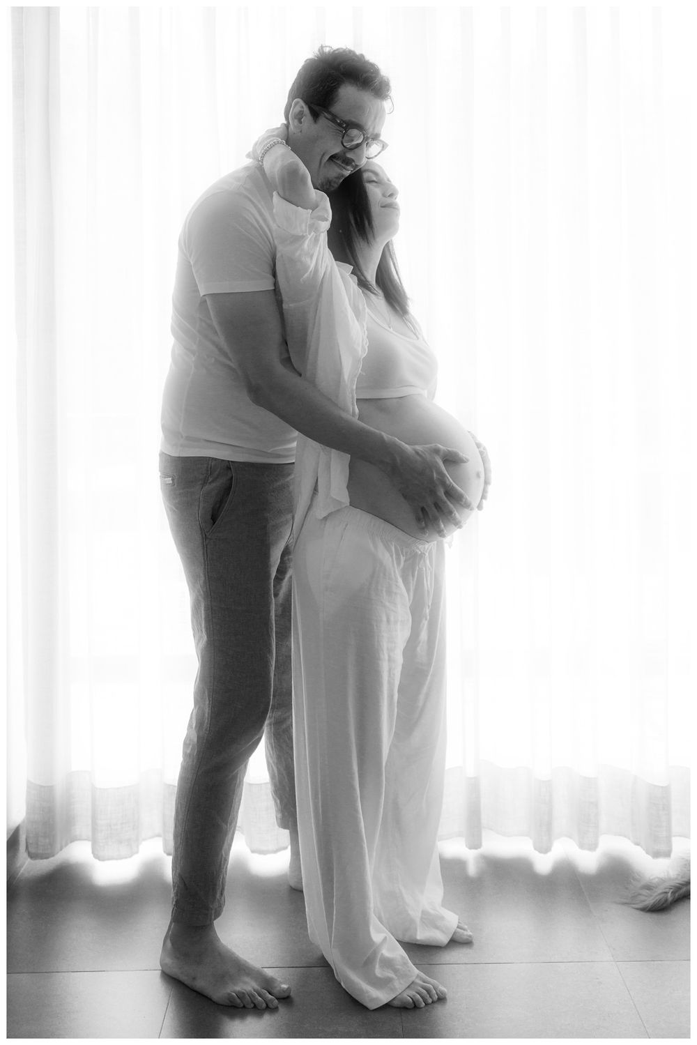 Black and white maternity photo of a couple embracing tenderly by a bright window, man cradling pregnant belly.