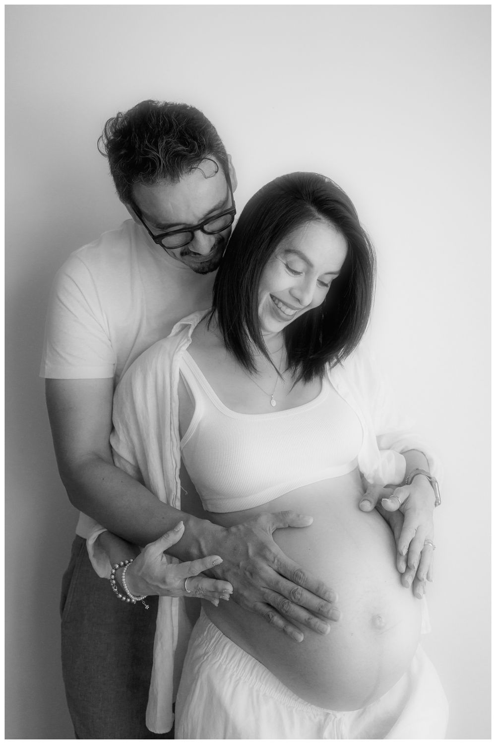 Black and white maternity photo of pregnant couple embracing, man holding woman's baby bump in studio portrait.