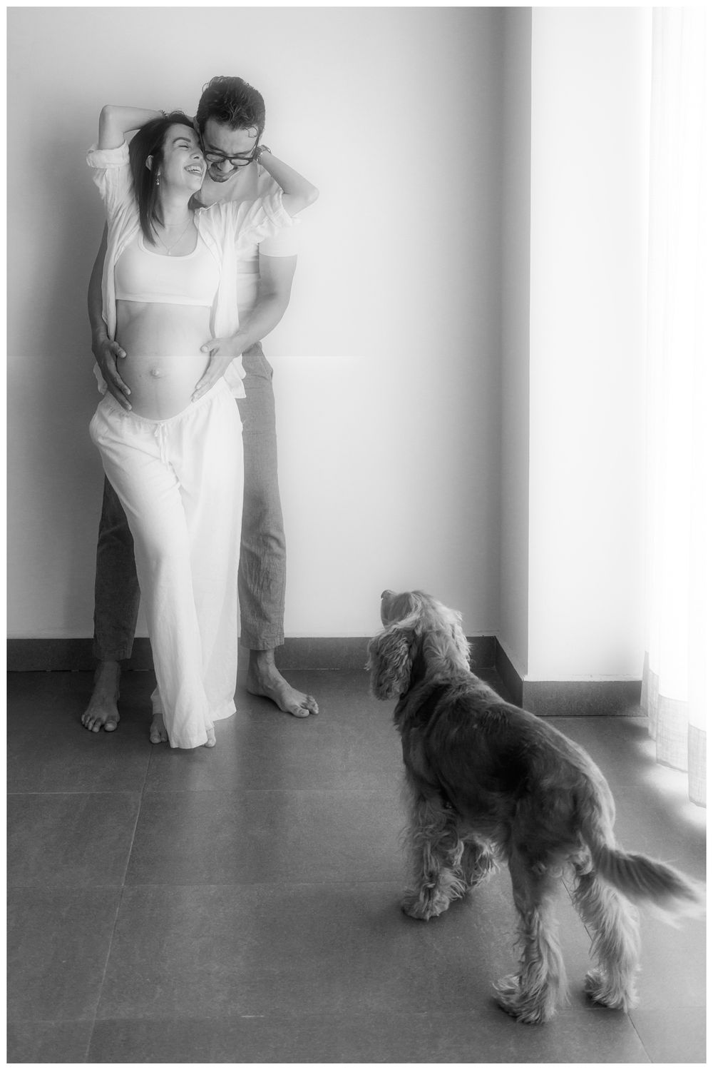 Pregnant couple embracing near window while dog looks up at them in black and white.
