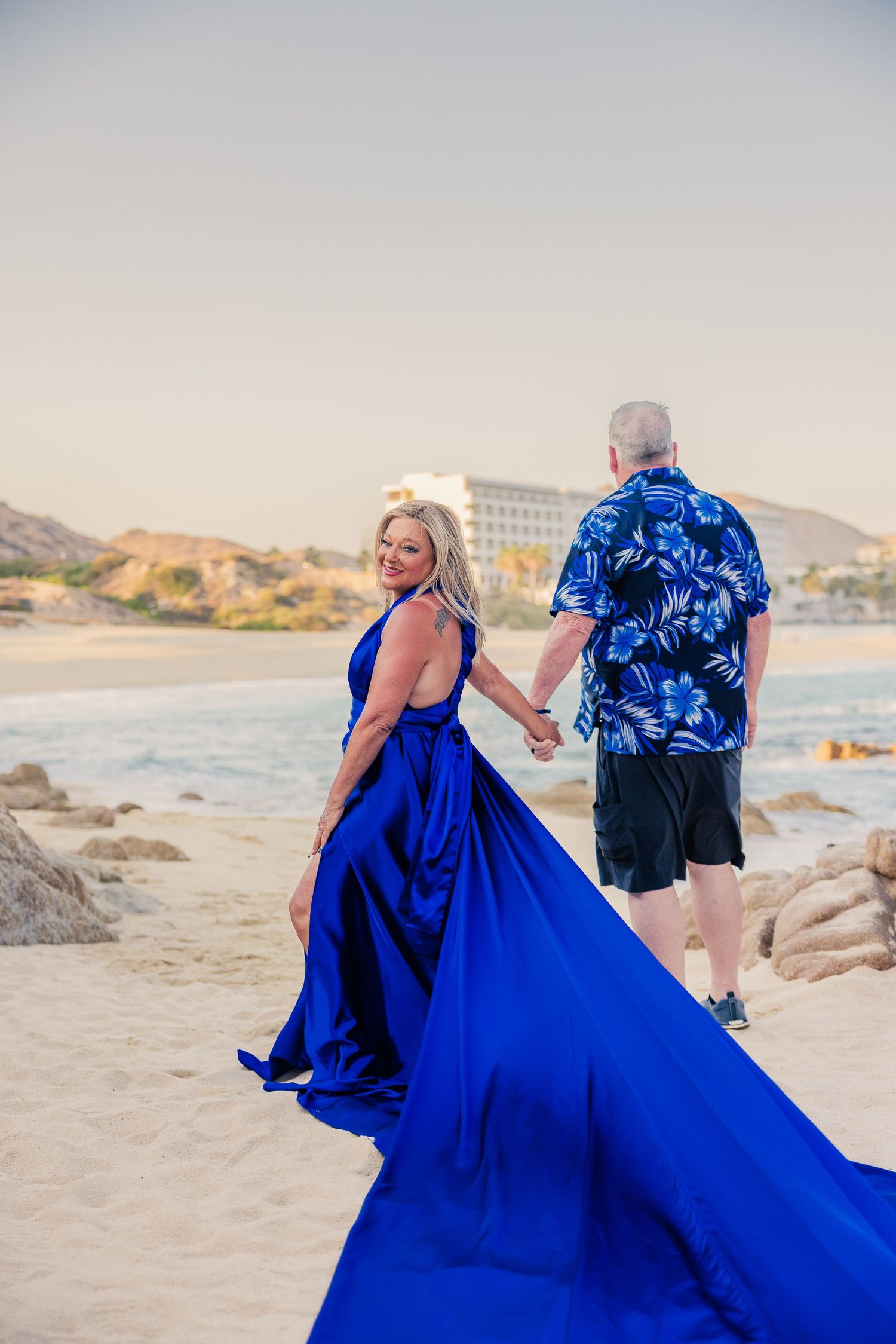 Couple walking on beach, woman in royal blue flowing dress smiling.