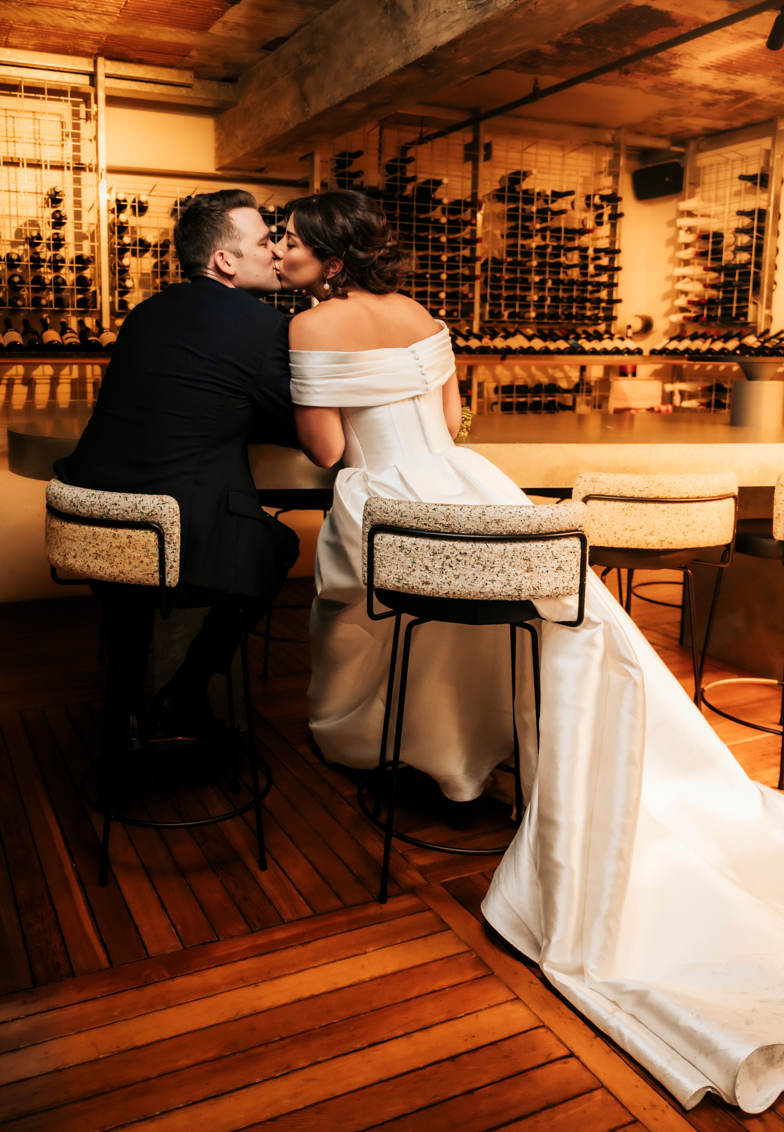 Newlyweds share a romantic kiss in an elegant wine cellar bar, bride in off-shoulder gown with flowing train.