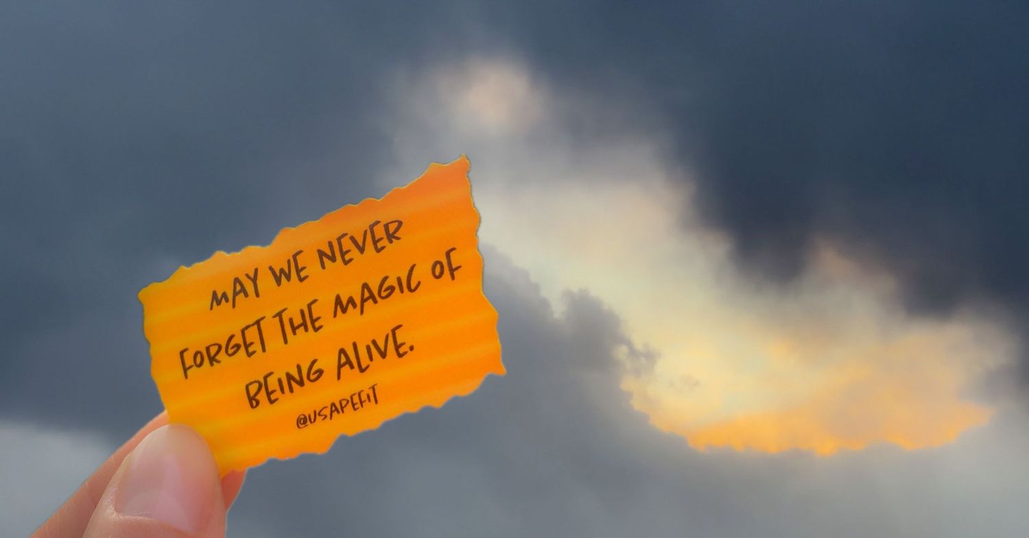 Hand holding an orange note card against a cloudy sunset sky with the quote May We Never Forget the Magic of Being Alive.