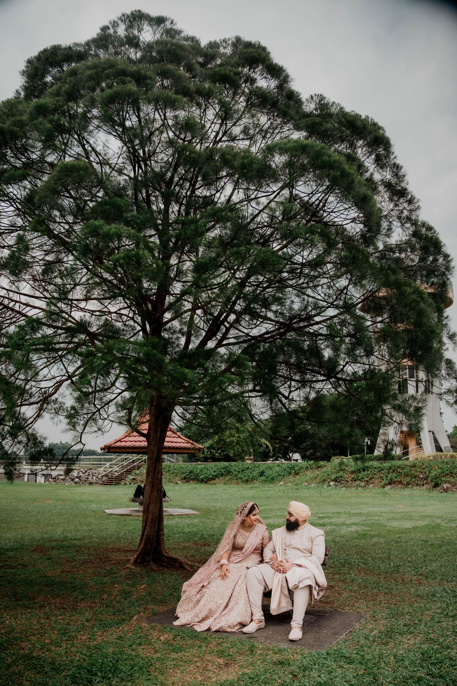 Couple in wedding attire resting under large trees in a peaceful park setting. Singapore