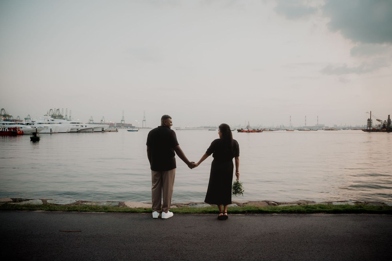 Silhouetted couple holding hands while standing at harbor during sunset with boats in background. Singapore