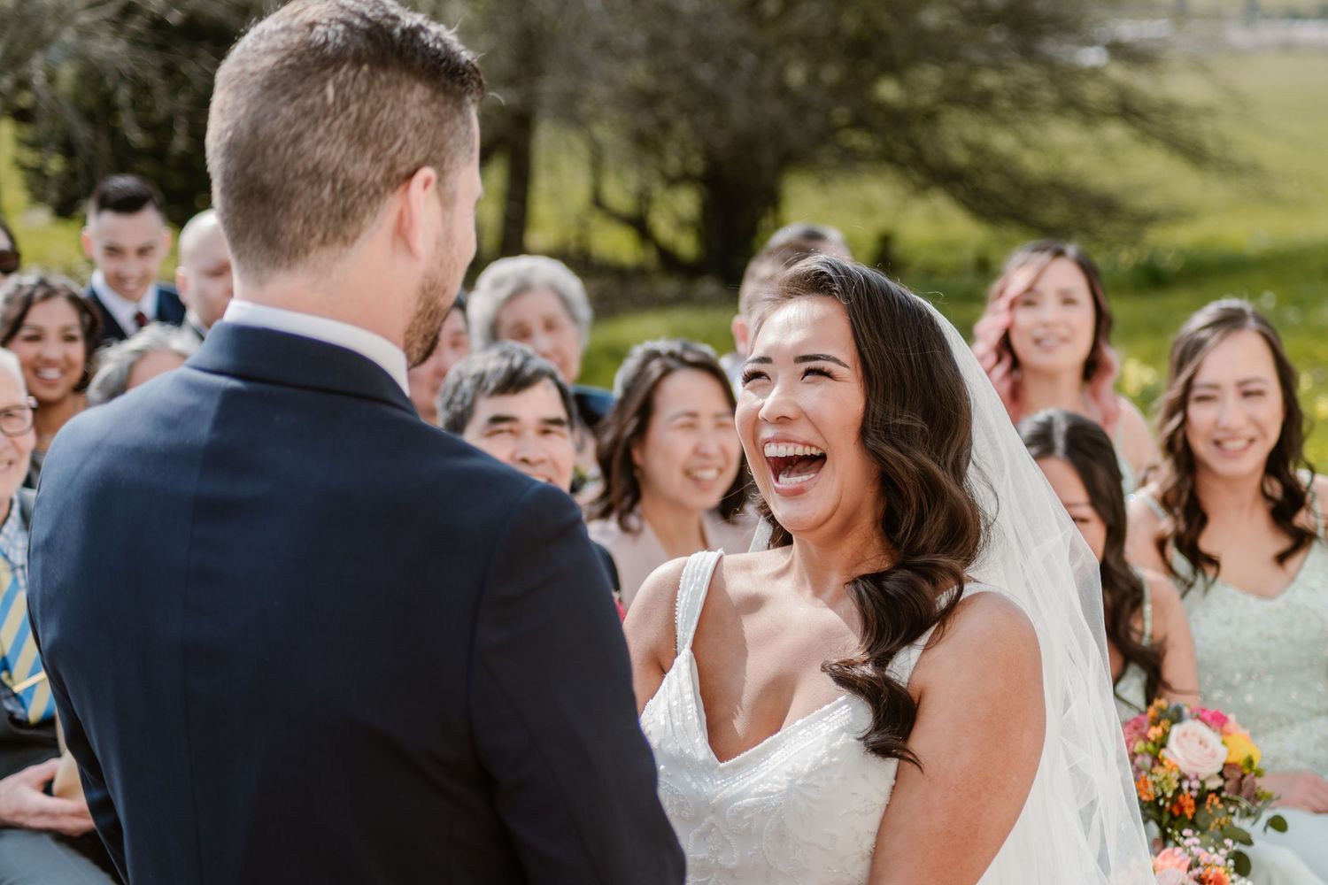 Bride and groom embrace in a lush garden, she laughs joyfully in a lace gown as he holds her in a blue suit.