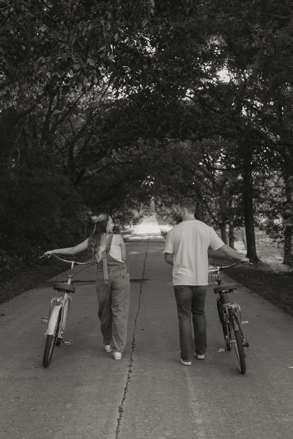 Black and white photo of a couple walking bicycles down a tree-lined path away from camera.