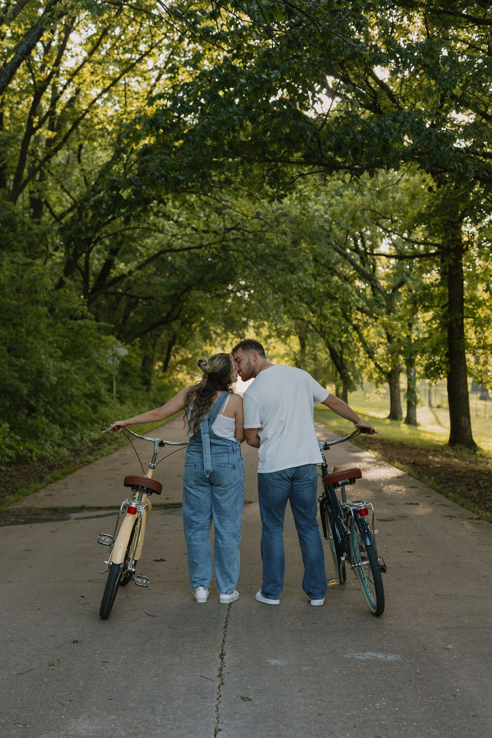 A couple stands on a tree-lined path with their bicycles, sharing a romantic moment in a lush green park.