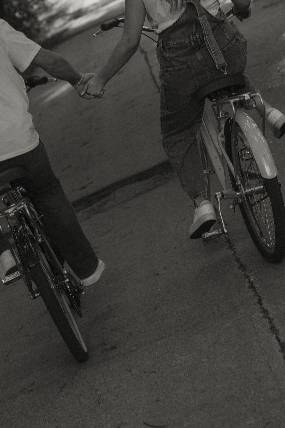 Black and white photo of a couple holding hands while riding bicycles together on a road.