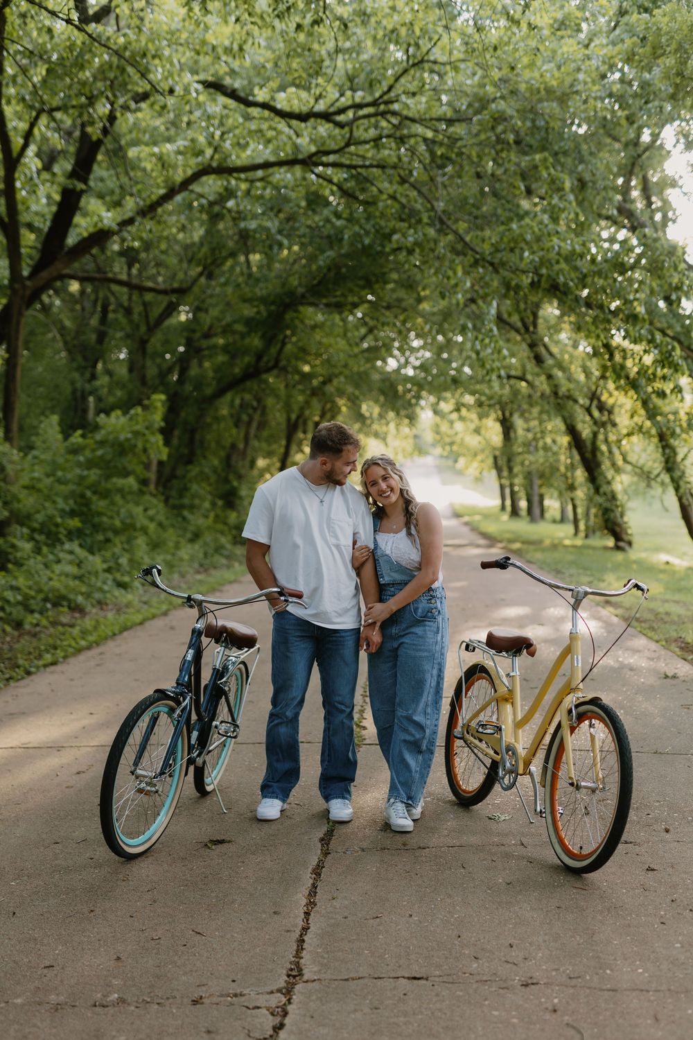 A couple stands together on a tree-lined path with two vintage bicycles, smiling and holding hands outdoors.