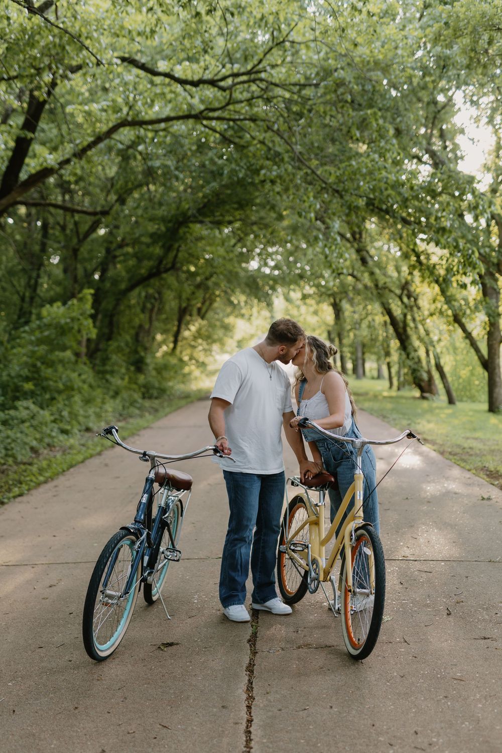Couple sharing a romantic moment between two vintage cruiser bicycles on a tree-lined path in a lush green park.