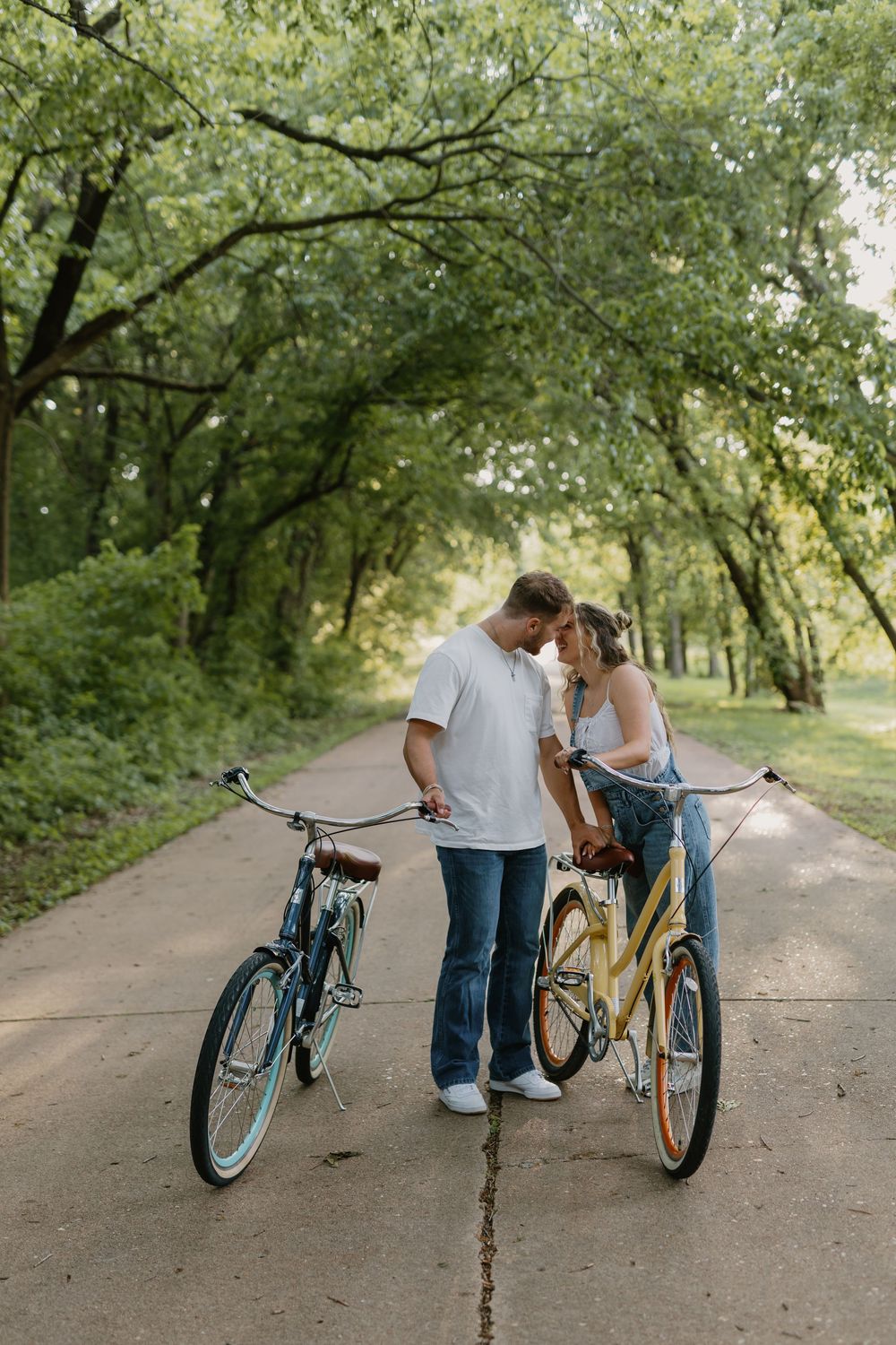 Couple sharing a romantic moment on a tree-lined path with vintage cruiser bicycles in a lush green park.