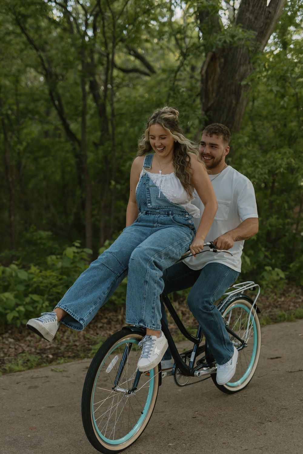 Couple riding a vintage bicycle with teal wheels through a lush green park, woman in denim overalls sitting on handlebars laughing.
