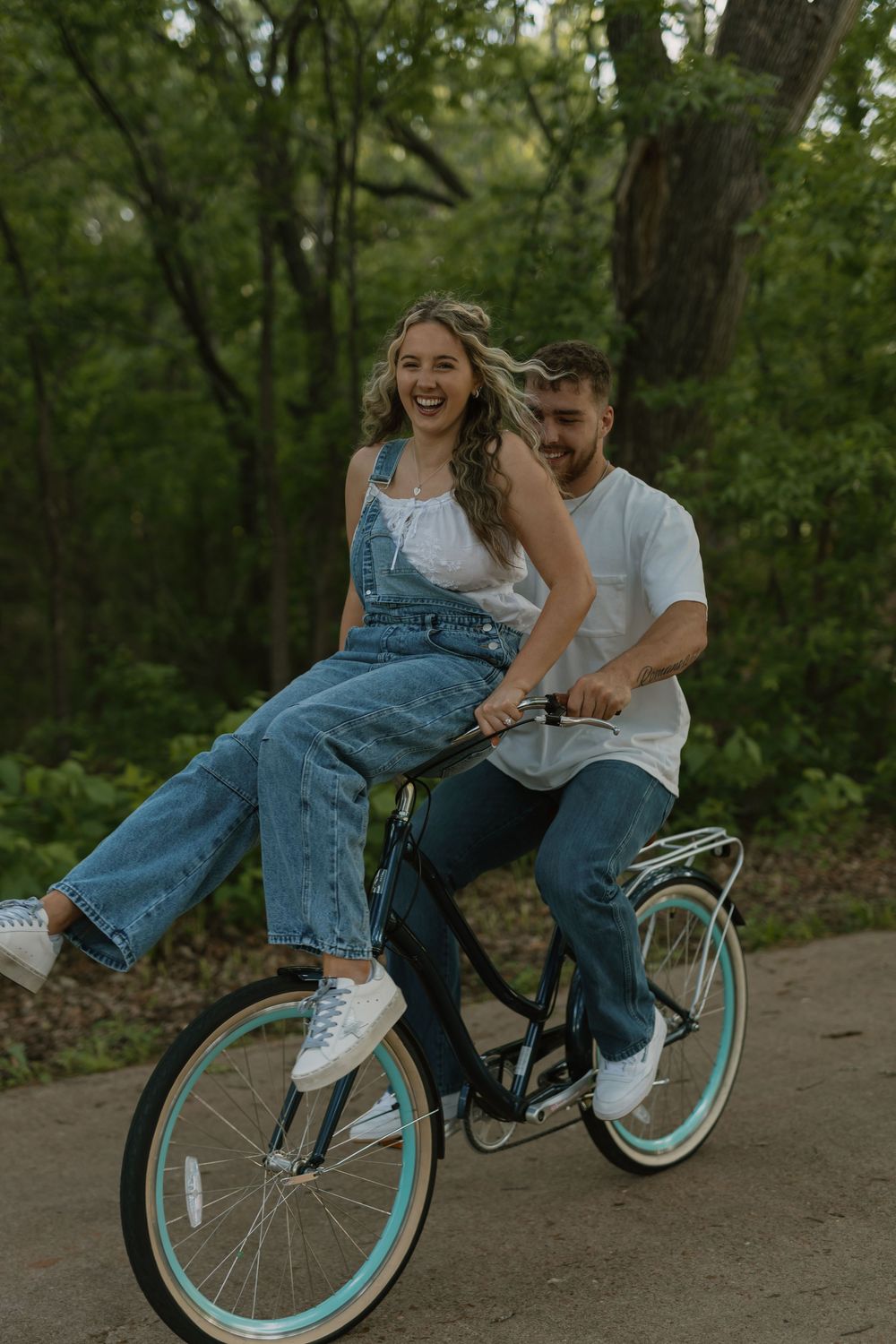 Happy couple sharing a vintage teal-wheeled bicycle on a tree-lined path, woman in denim overalls laughing joyfully.