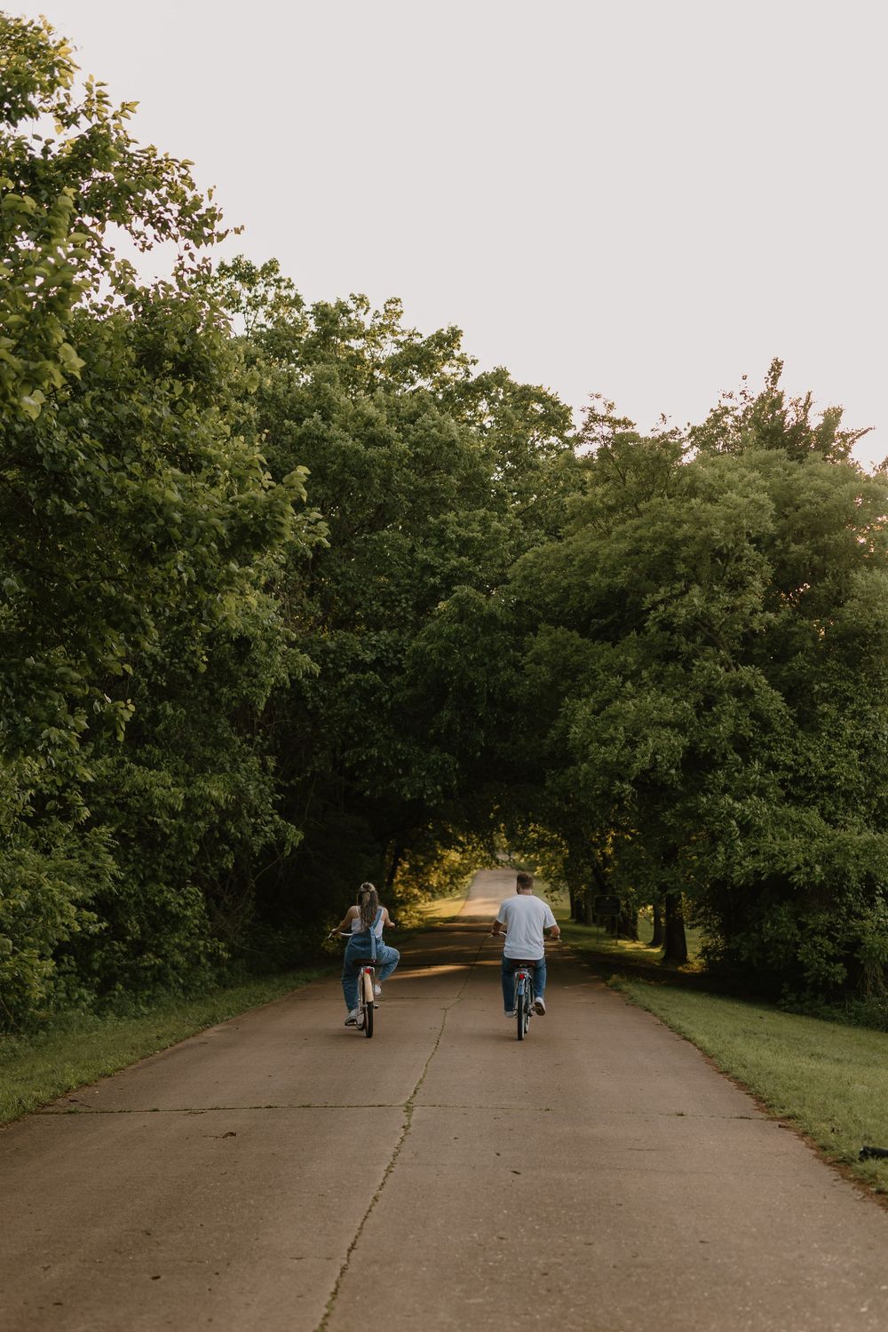 Couple riding bicycles together down a scenic tree-lined path at sunset.