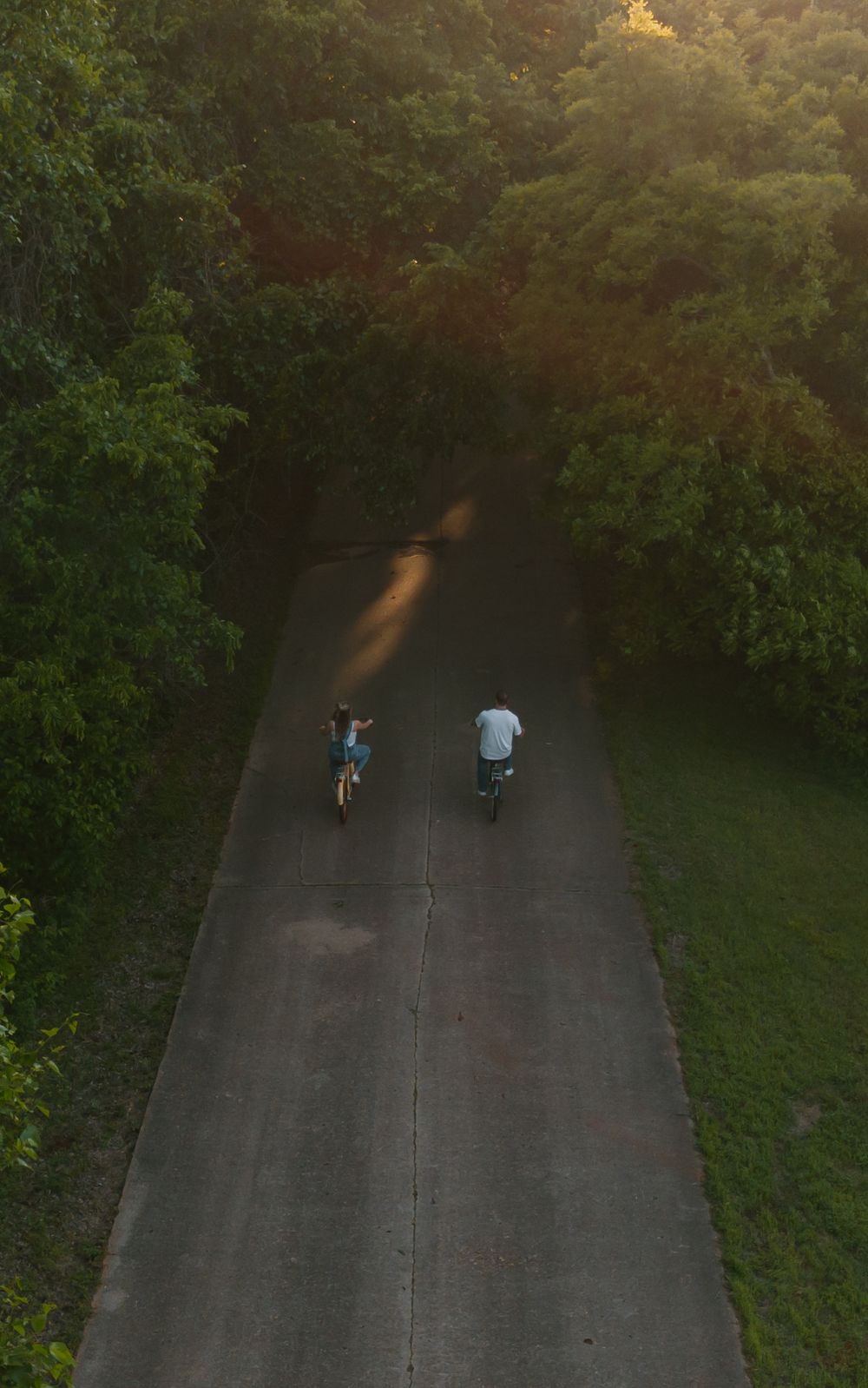 Aerial view of two cyclists riding bikes on a narrow tree-lined road bathed in warm golden sunset light.