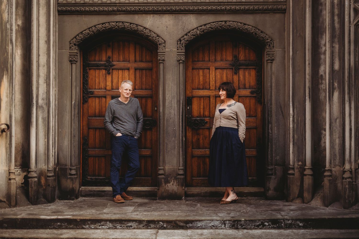 A man and woman stand apart before ornate wooden doors of a historic stone building.