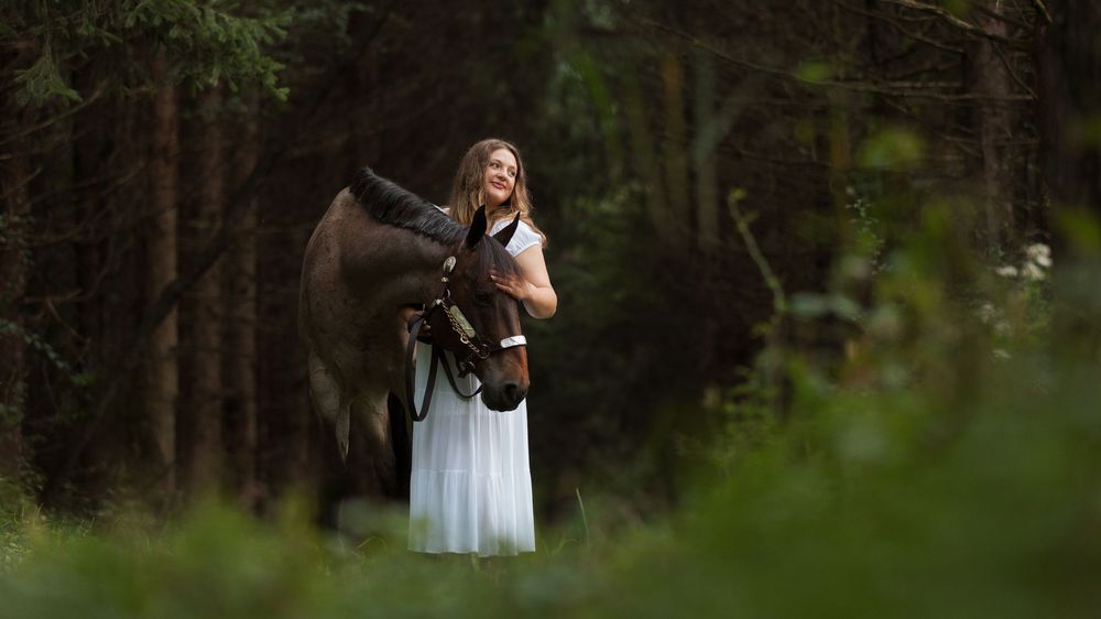 Woman in white dress standing with a dark horse in a lush green forest setting.
