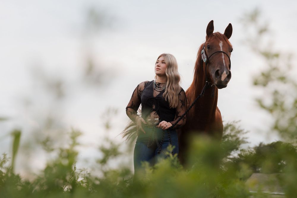 Young blonde woman standing beside a chestnut horse in a lush green field at dusk.
