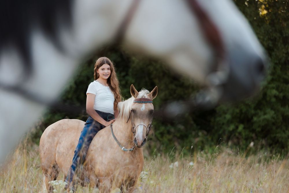 A young girl rides a palomino horse in a grassy field, with another horse in the foreground.