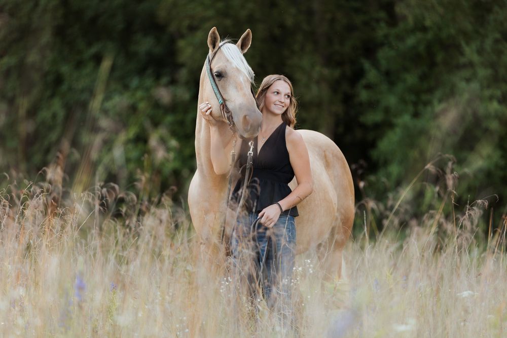 Woman standing with a palomino horse in a golden meadow with lush green trees in the background.