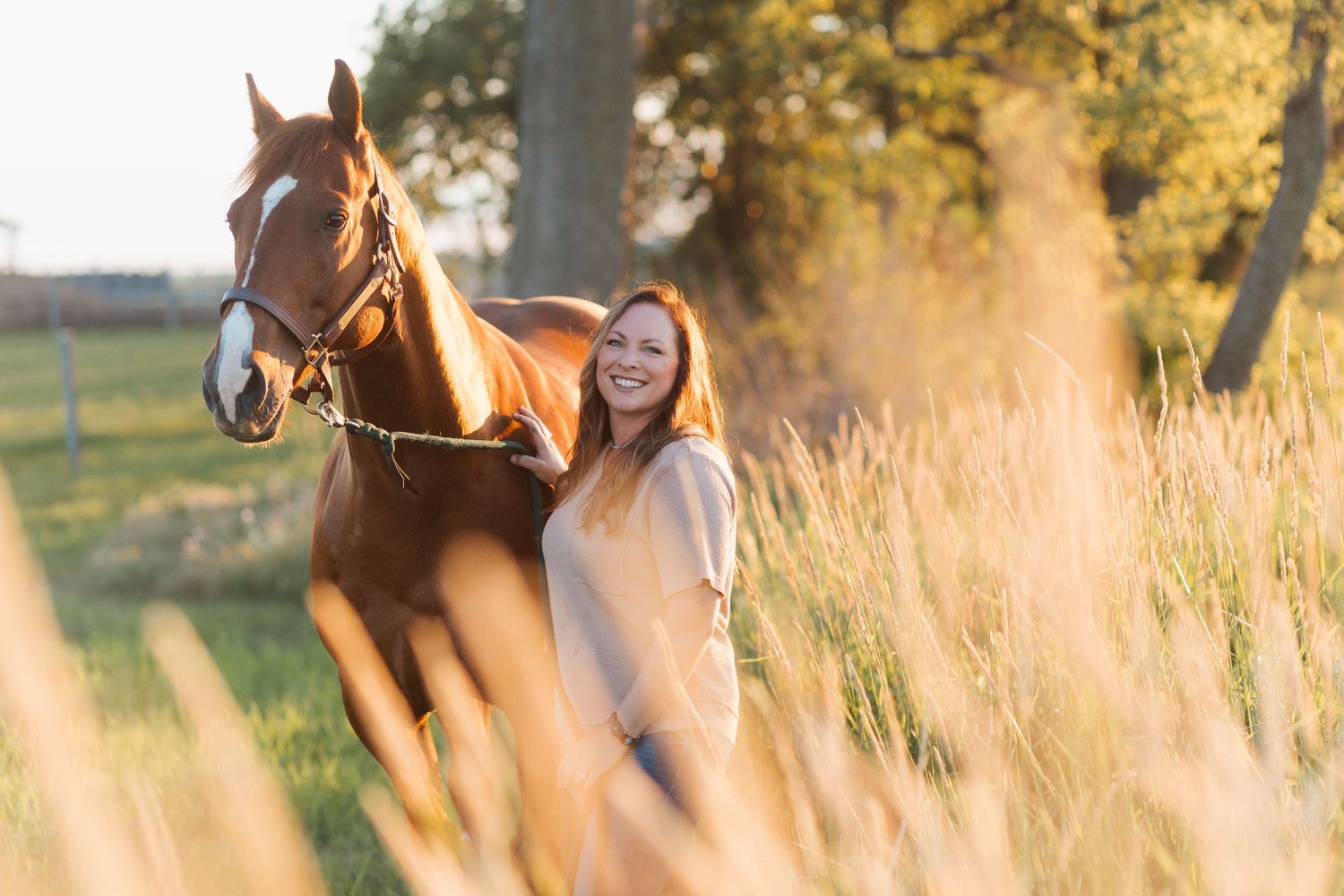 Smiling redhead woman standing beside a chestnut horse in a sunlit golden field with tall grass and trees.