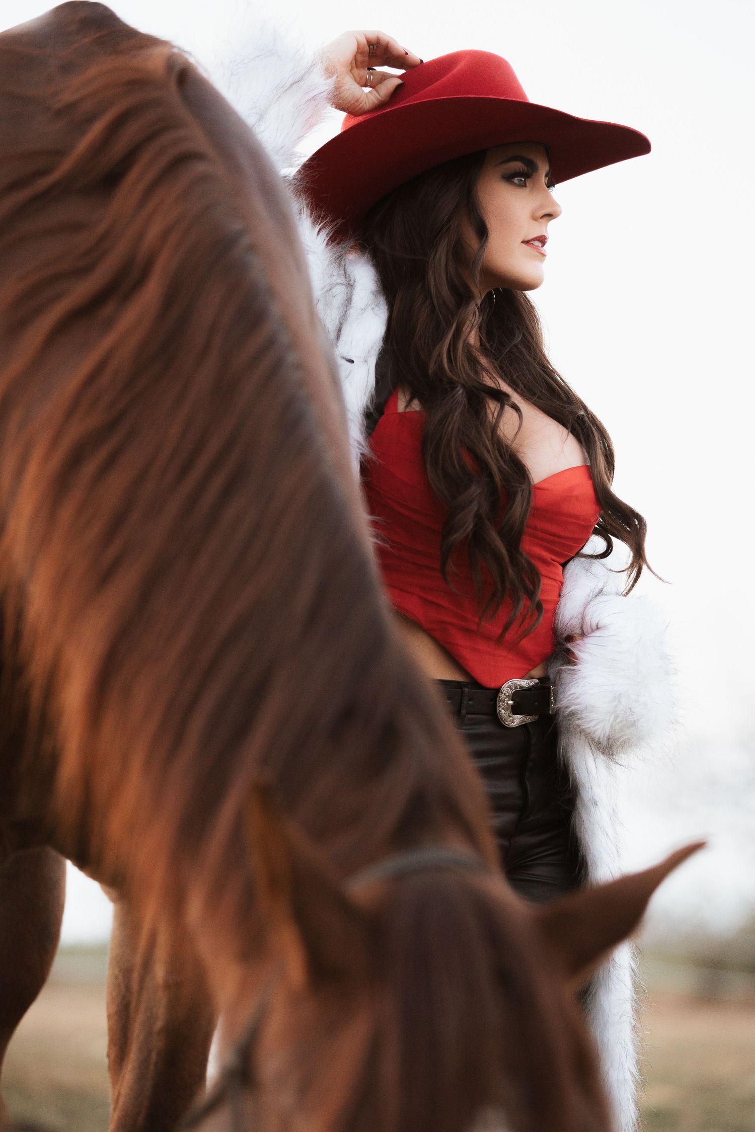 Woman in red hat and coat stands beside a majestic brown horse in a dreamy winter setting.