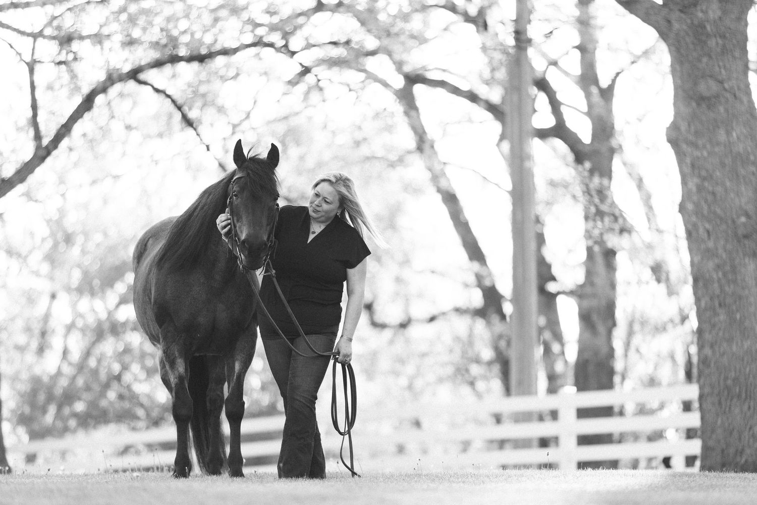 Black and white photo of a woman standing with a dark horse on a lead rope near a white fence and bare trees.
