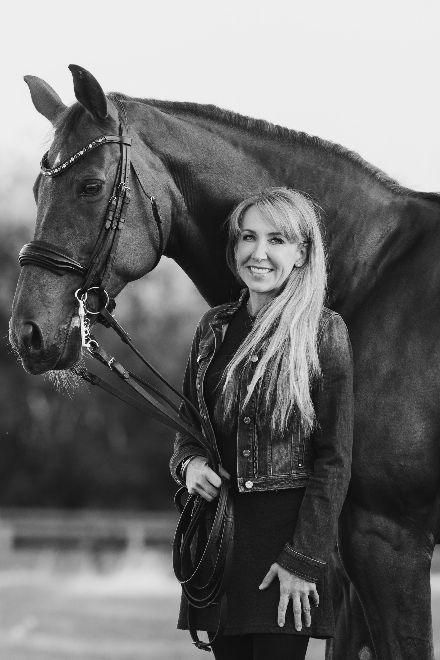 A black and white portrait of a woman in dark clothing standing beside a majestic dark horse in equestrian gear.