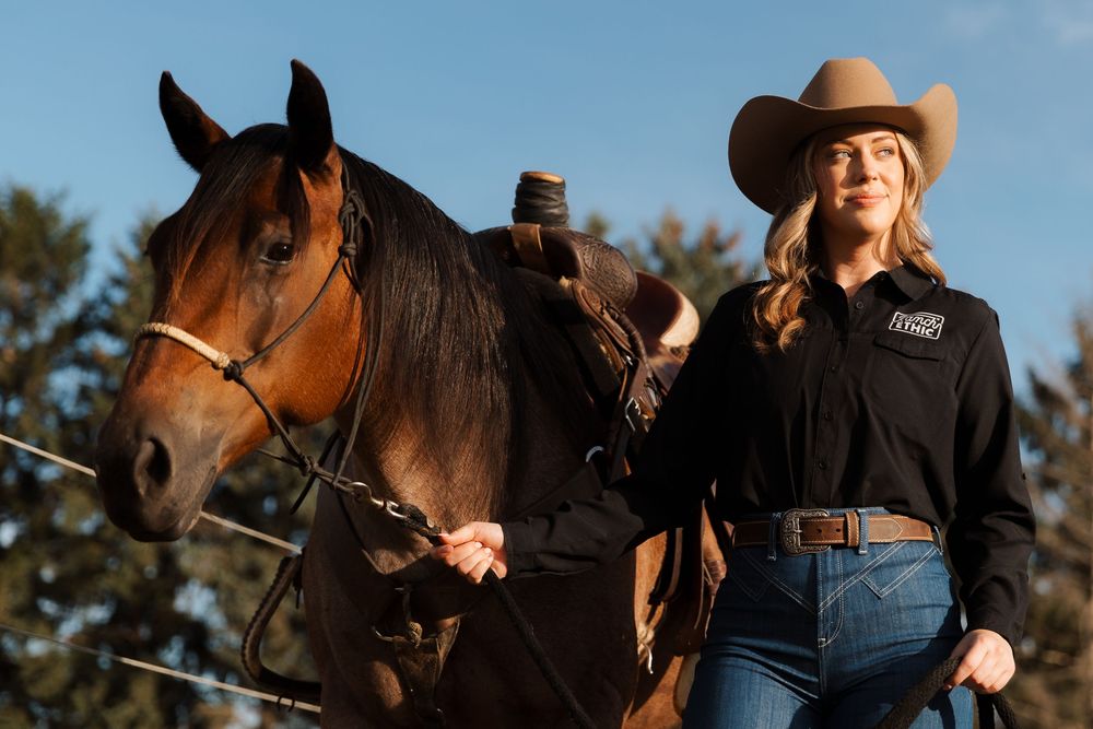 A blonde woman in a cowboy hat and black shirt stands beside a bay horse outdoors in golden hour light.