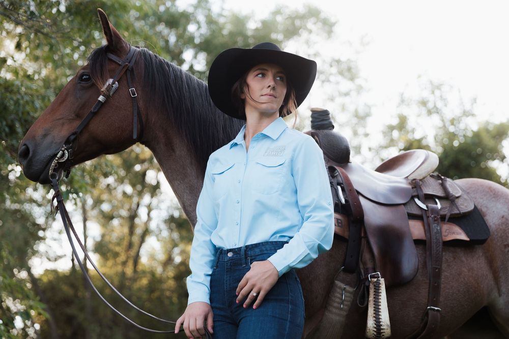 Woman in cowboy hat and blue shirt standing beside a saddled brown horse outdoors.