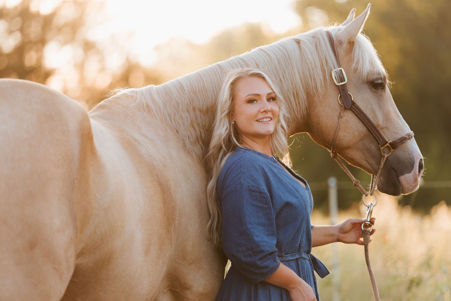 Young blonde woman in blue dress smiling beside a palomino horse in a sunny golden field.