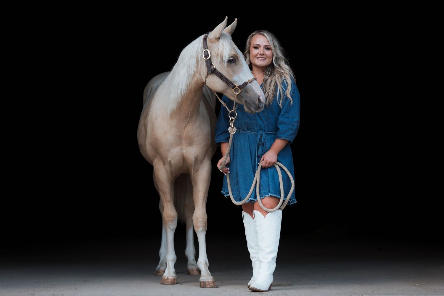 Blonde woman in blue dress and white boots poses with a white horse against a dark studio background.