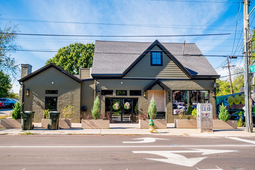 Dark green commercial building with black roof, large windows, and wraparound porch on a sunny street corner.