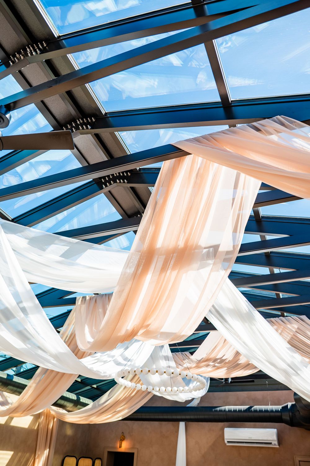 Elegant wedding venue ceiling draped with flowing white and blush fabric under a glass skylight.