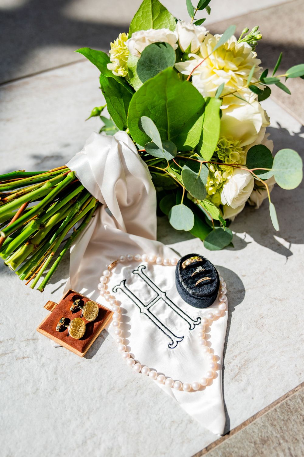Wedding flat lay with white rose bouquet, pearl necklace, gold cufflinks, rings, and monogram handkerchief.