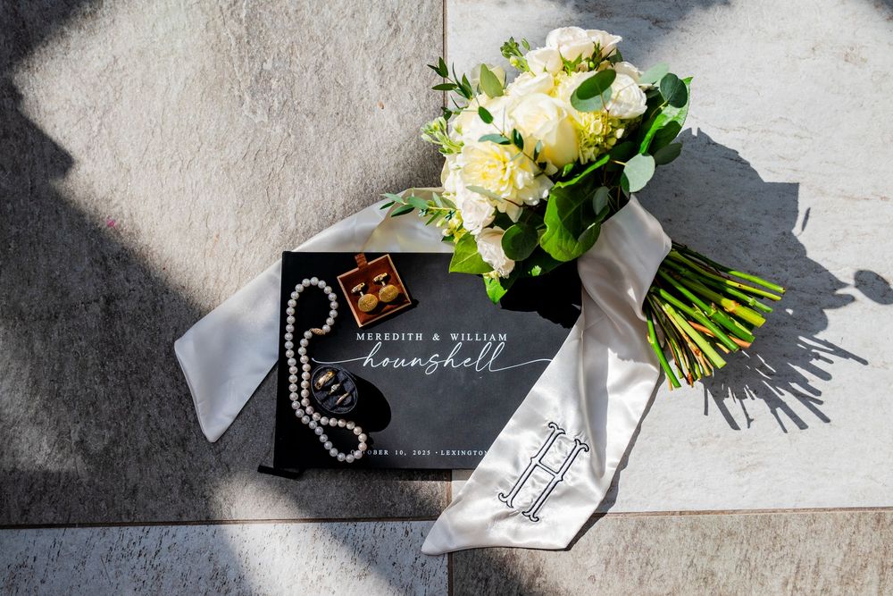 Wedding details flat lay featuring Hounshell wedding book, white bouquet, pearl necklace, and gold cufflinks.