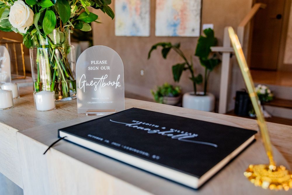 Elegant wedding guestbook display with black hardcover book and acrylic sign on white table with floral decor.