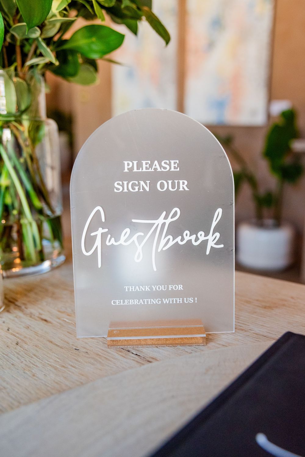 Elegant frosted acrylic arch wedding guestbook sign on a wooden stand with greenery backdrop.