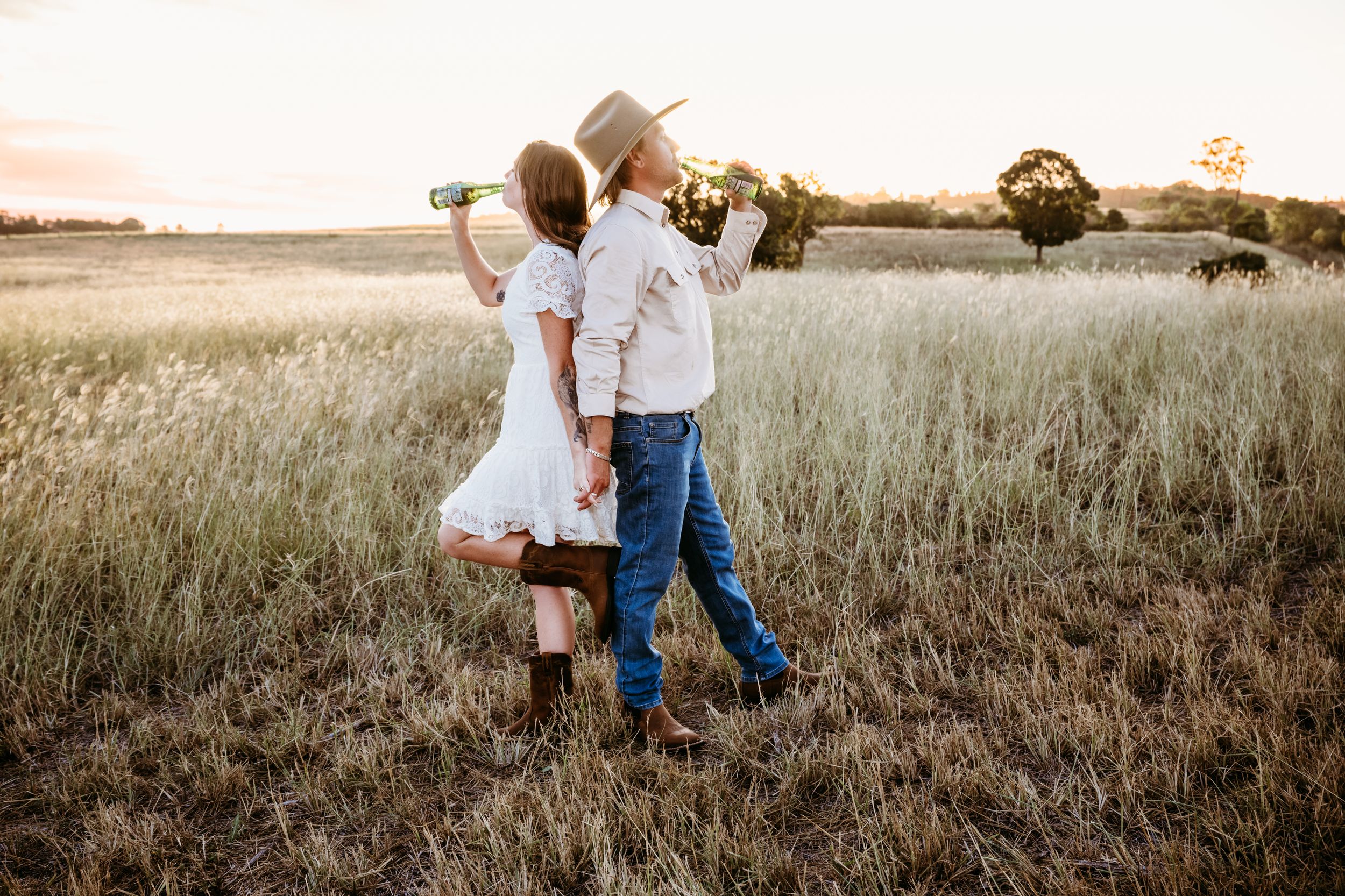 Couple walking hand in hand through golden field at sunset, woman in white dress, man in cowboy hat and jeans.