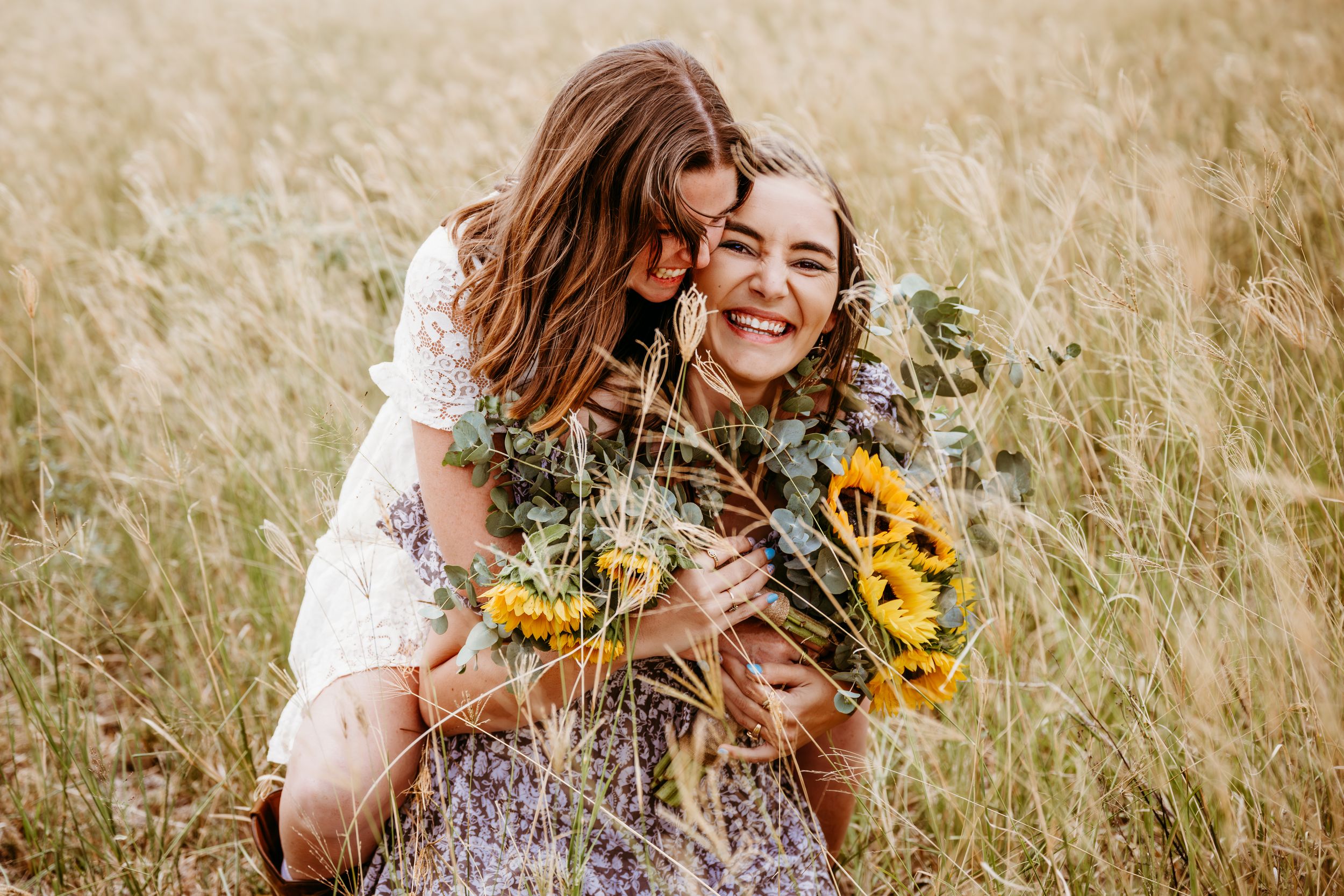 Two young women embrace joyfully in a golden field holding sunflowers.